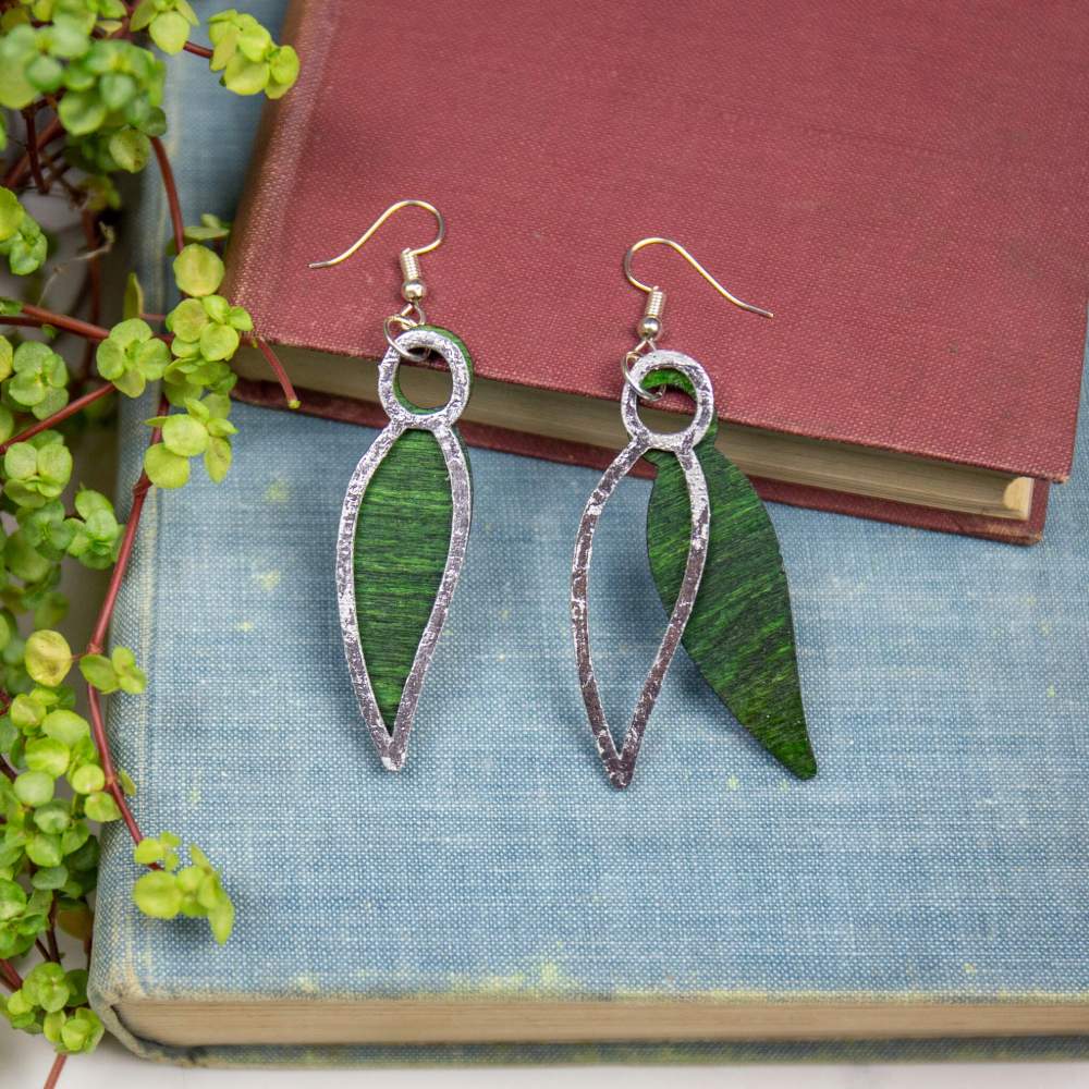 A pair of wooden green leaf-shaped earrings with a silver accent layer, displayed on 2 stacked books.