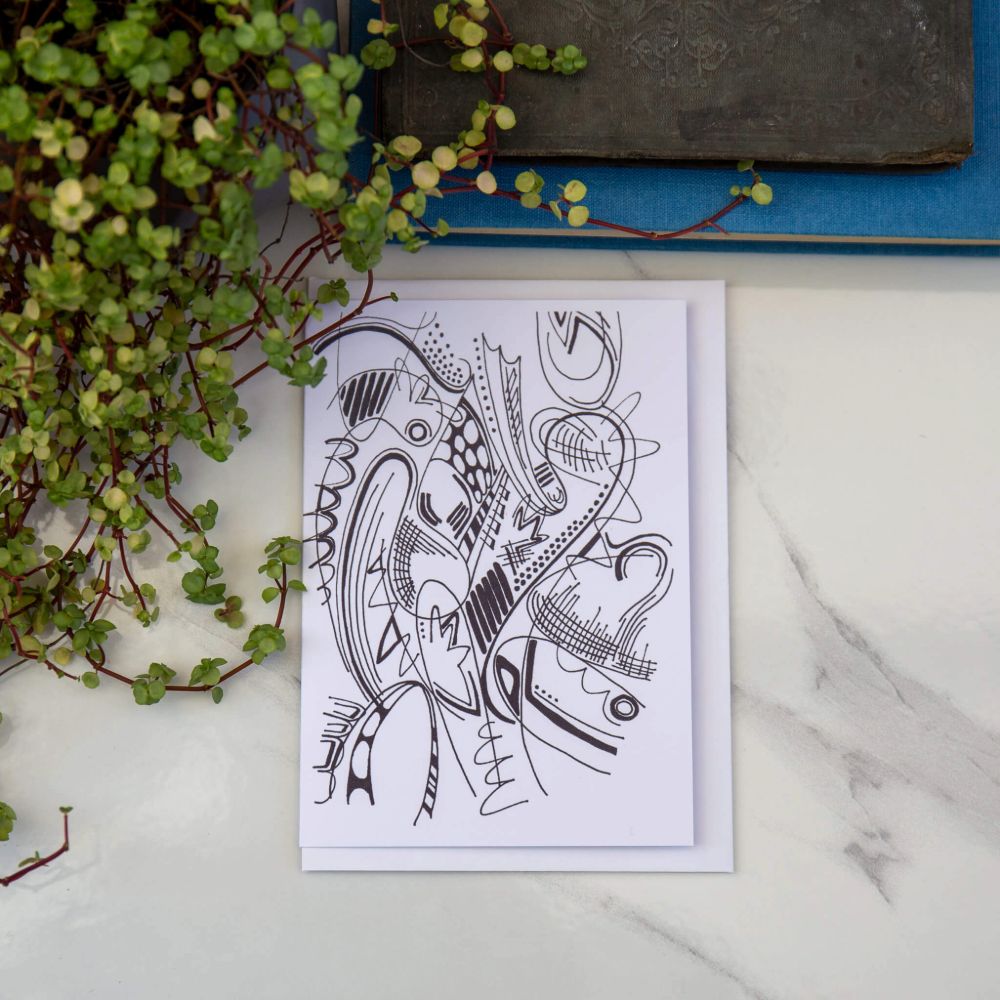 A greetings card with an abstract black and white ink illustration, placed on a marble surface with a blue book and a potted plant in the background.