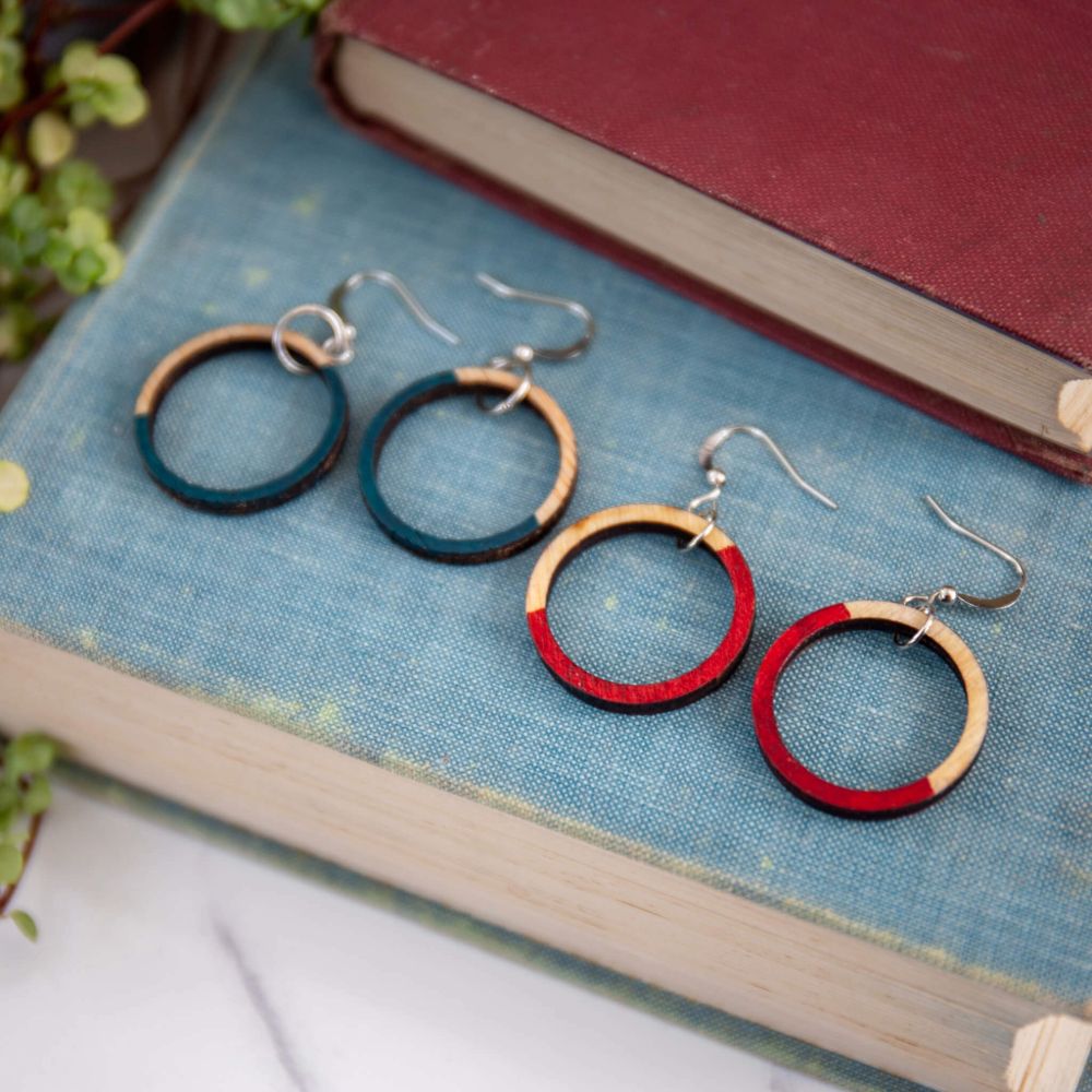2 pairs of wooden circle earrings, with half the circles hand painted. One pair blue, one pair red, lying on a blue clothbound book.