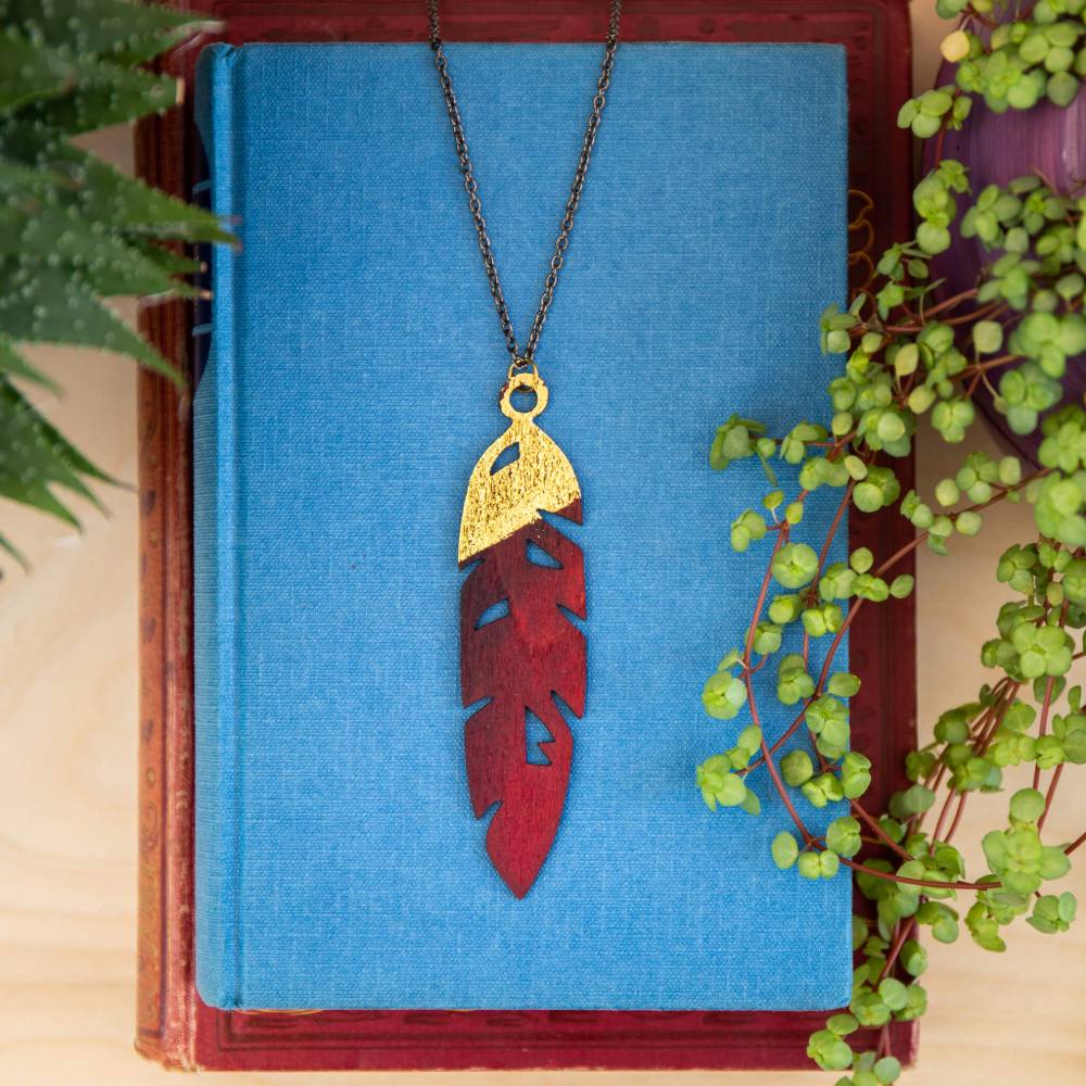 Deep red hand painted wooden feather necklace with gold foiling detail laid on blue book.