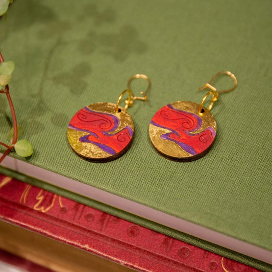 2 wooden gold and red swirling planet earrings laid on a green clothbound book.