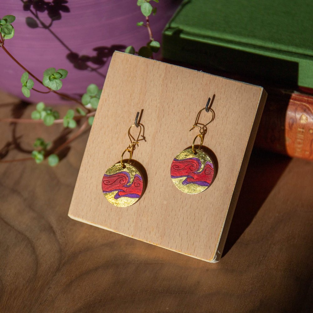 A pair of red and purple swirling planet earrings with gold foil details, hanging on a wooden stand.