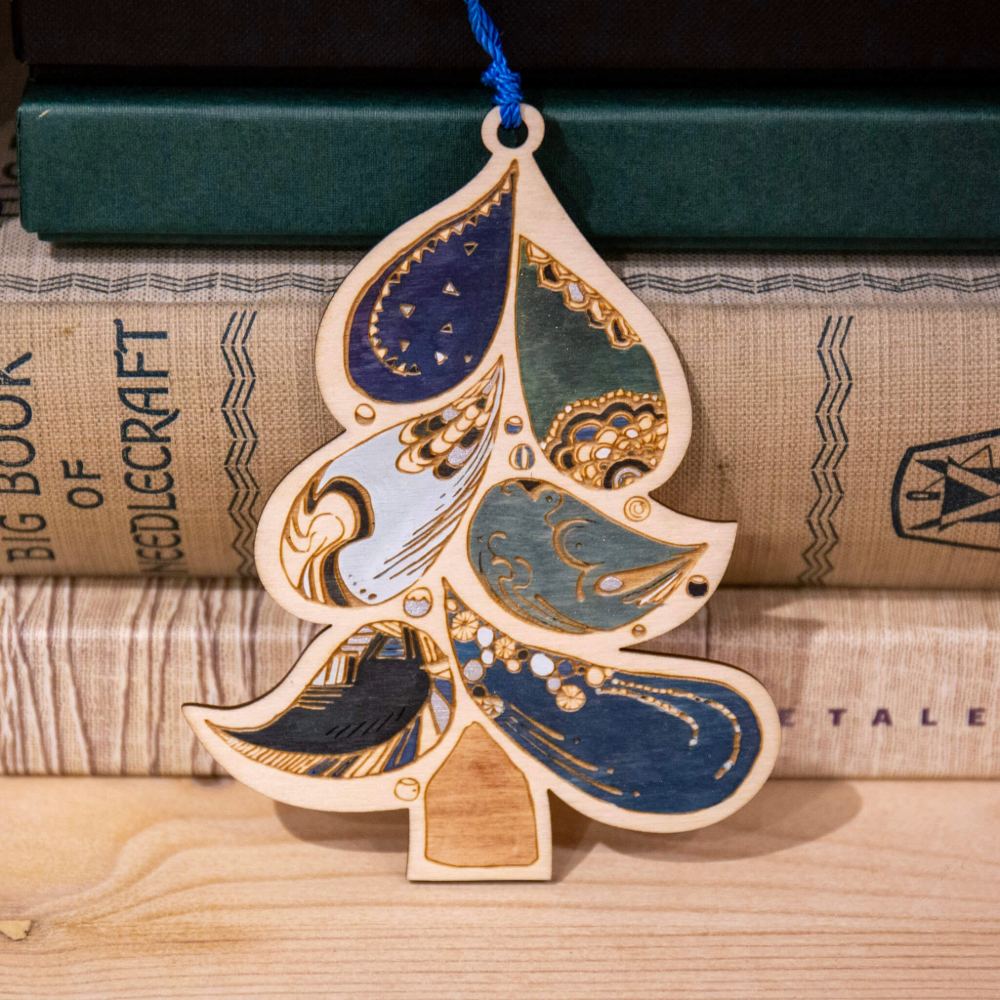 An engraved and hand-painted, paisley-inspired Christmas tree decoration, in blues and grey, displayed against a stack of vintage books.