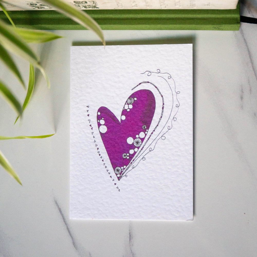 A textured greetings card featuring a purple illustrated heart design featuring swirling lines and circle patterns, laid on a white marble surface.