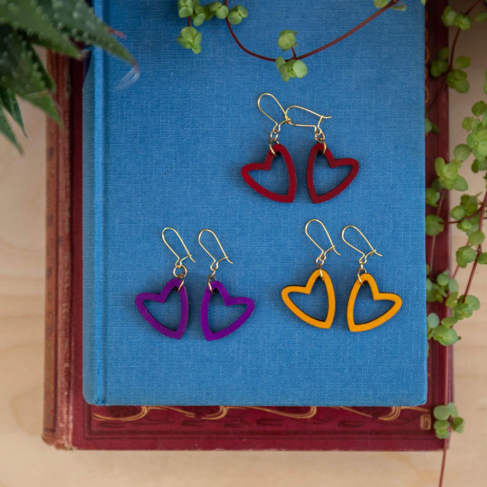 3 heart shaped earrings, in red, yellow and purple, laid on a blue clothbound book.