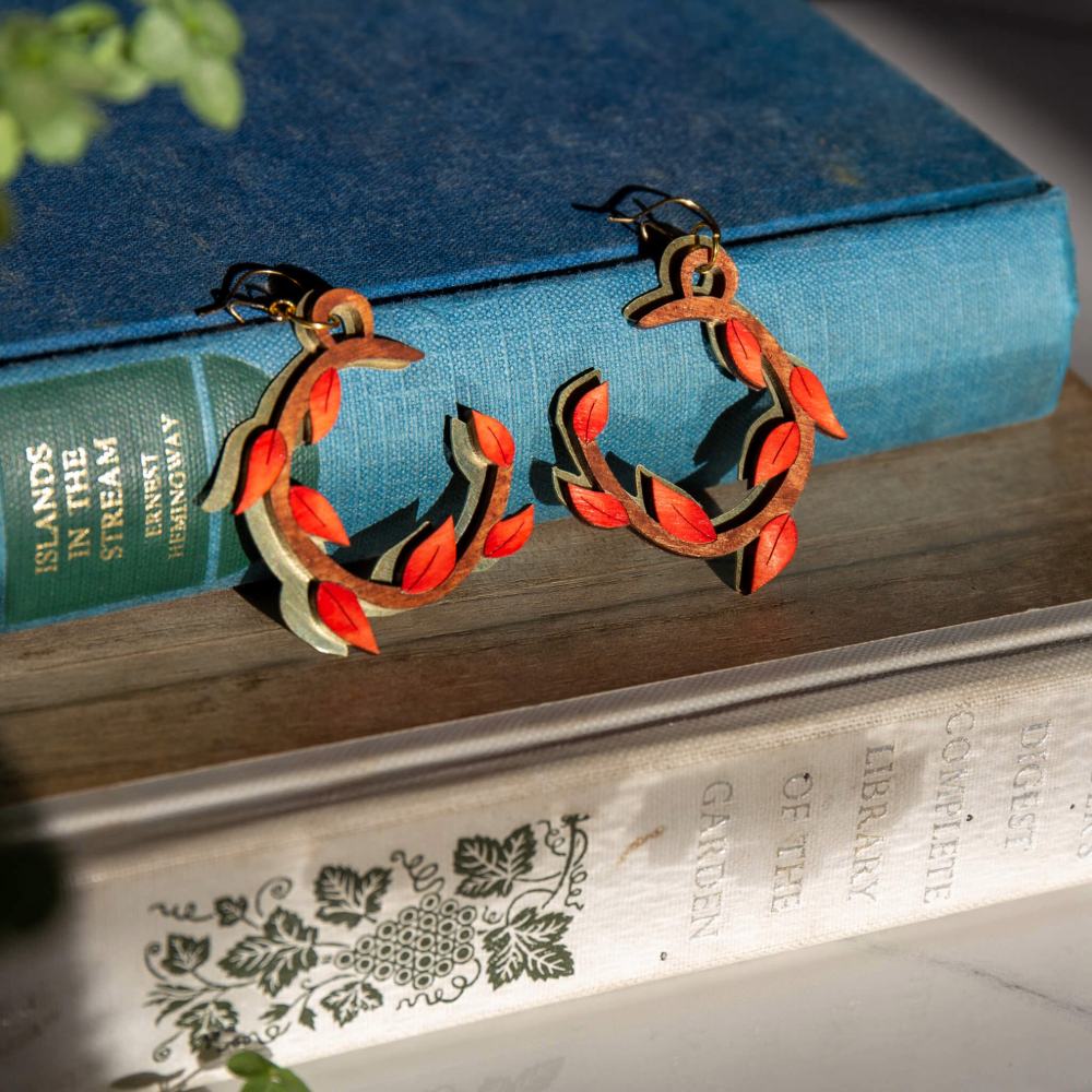 A pair of leafy hoop earrings in rich burnt orange layered over a matching wreath of hand cut brass, leaning against a pair of clothbound books.