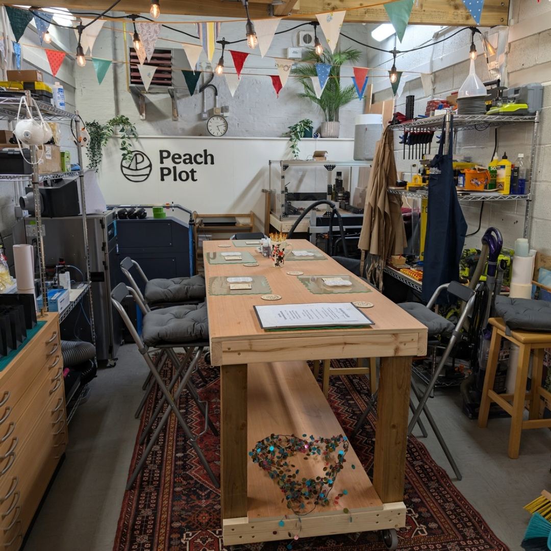Workshop & maker space with a table, chairs, and various tools and equipment. Peach Plot logo visible on the wall.