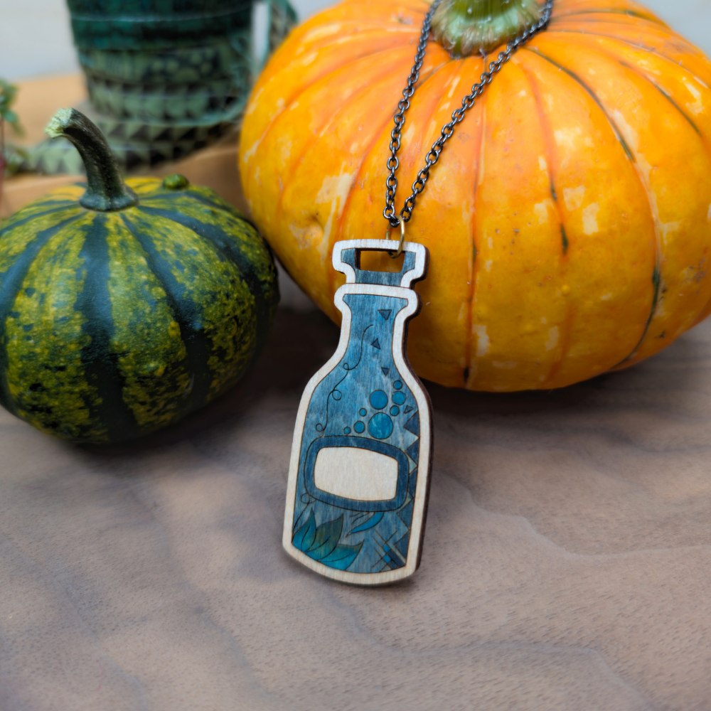A wooden potion bottle necklace with hand-painted details in greys and blues. displayed on miniature pumpkins with a vintage-style chain.
