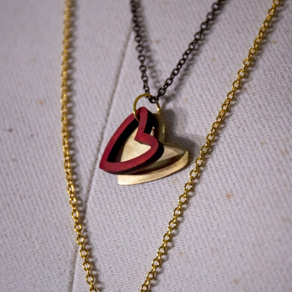 A necklace with a red wooden heart pendant layered over a gold-coloured hand-cut brass heart, hanging on an vintage style chain.