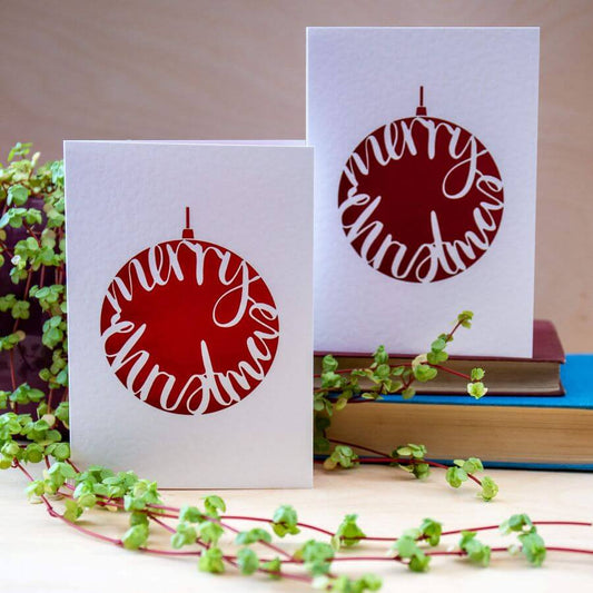 Two Christmas-themed greeting cards with red and white designs on a surface with greenery and books.