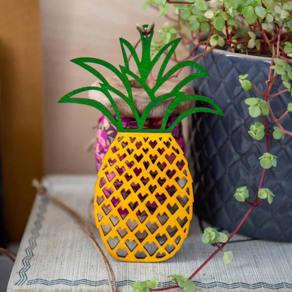 A hand-painted, laser cut wooden pineapple decoration with a lattice of tiny hearts, in vibrant yellow and green colors, placed on a shelf.